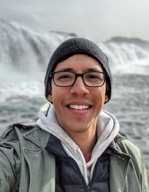 Allen Schober smiling while wearing black rimmed glasses, a green jacket, and a black beanie in front of a waterfall.